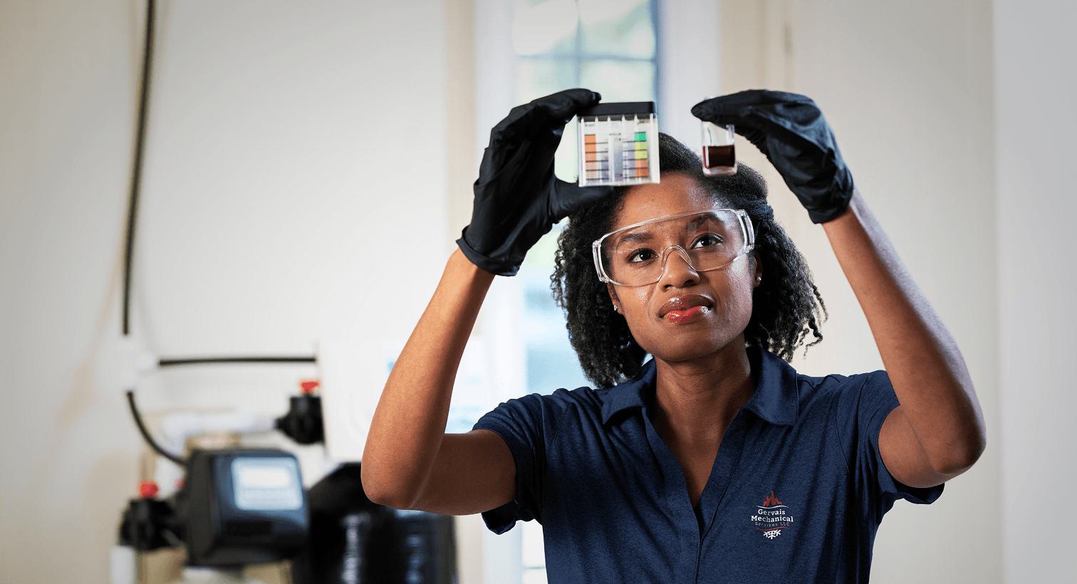 A Gervais technician in a blue uniform and safety gear compares a water sample to a testing chart in front of a water softener system.
