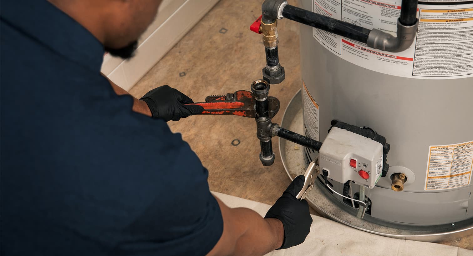 A Gervais plumber wearing black gloves uses a pipe wrench to repair the gas line and control valve assembly on a residential water heater tank.